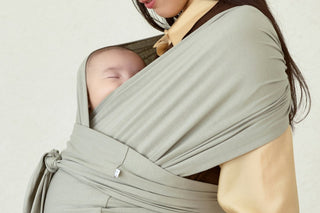 Sleeping baby in a sage green Konny Baby Carrier FLEX SoftBreeze™ worn over a yellow shirt, plain wall