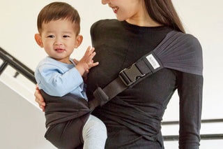 Smiling toddler seated in a black Dual Support Hip Seat Carrier worn by a woman, stair railing behind