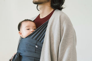 Baby looking out from a navy Konny Baby Carrier FLEX worn by a man in a grey cardigan, plain white wall