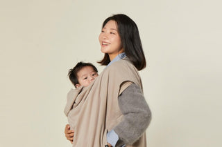 Smiling woman in a beige Konny carrier warmer with a baby peeking out at her chest, beige studio backdrop