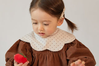 Toddler girl looking down in a Konny Bib Rolling in Wine Dot over a brown dress, holding a red toy, plain wall
