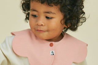 Curly-haired toddler looking down in a Salmon Konny Bib Rolling with a penguin label, over a cream top, beige wall