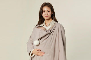 Smiling woman in a taupe Konny carrier warmer with a baby tucked inside at her chest, beige studio backdrop
